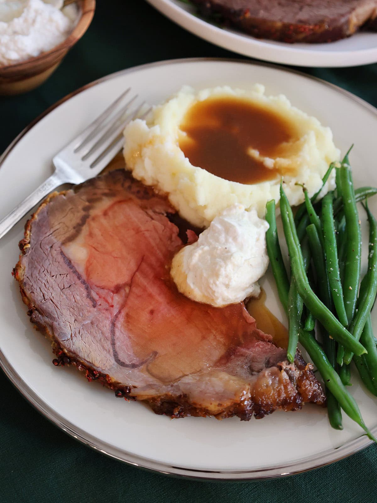 A slice of prime rib on a plate next to green beans, creamed horseradish, and mashed potatoes.