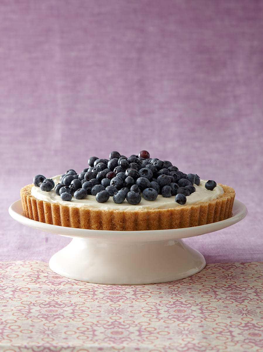 Side view of a simple no bake cheesecake topped with fresh blueberries, on a white cake stand. 