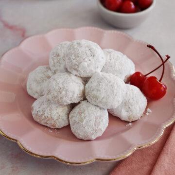 A pink plate piled with cherry almond cookies next to cherries.