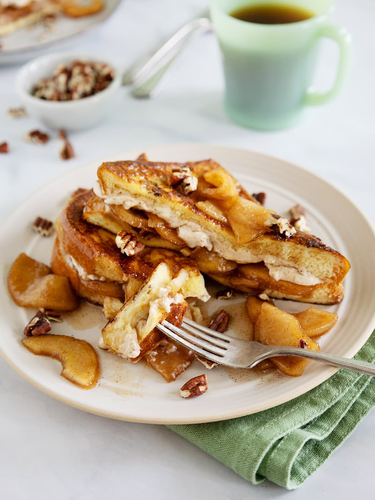 Apple French toast on a plate with a bite held up on a fork next to a green mug and green napkin.