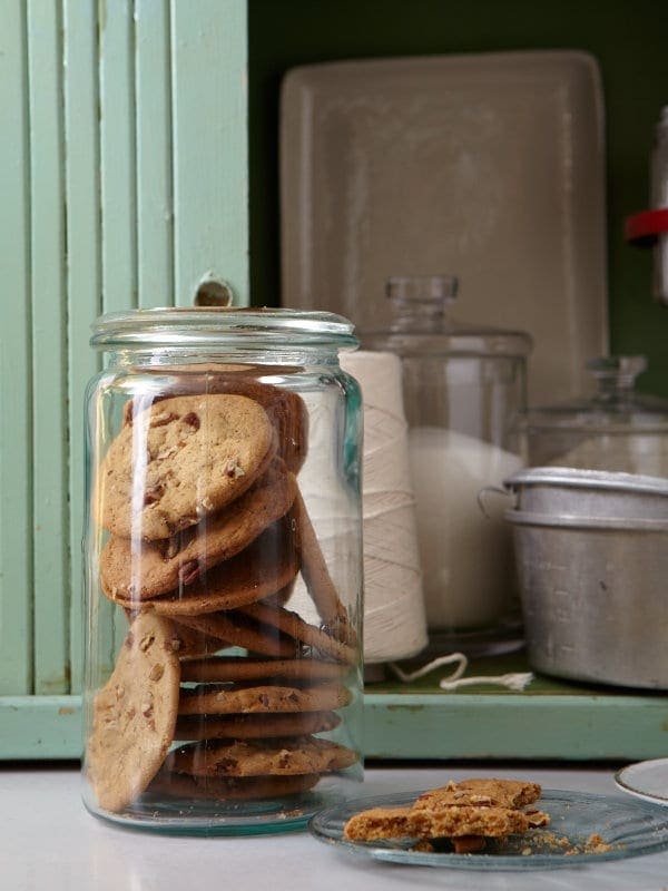 A glass jar full of crispy pecan cookies. A small plate lies near the jar, wit ha half eaten cookie on it. 