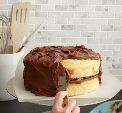 Classic yellow cake with chocolate frosting on white cake plate - process is cake being frosted
