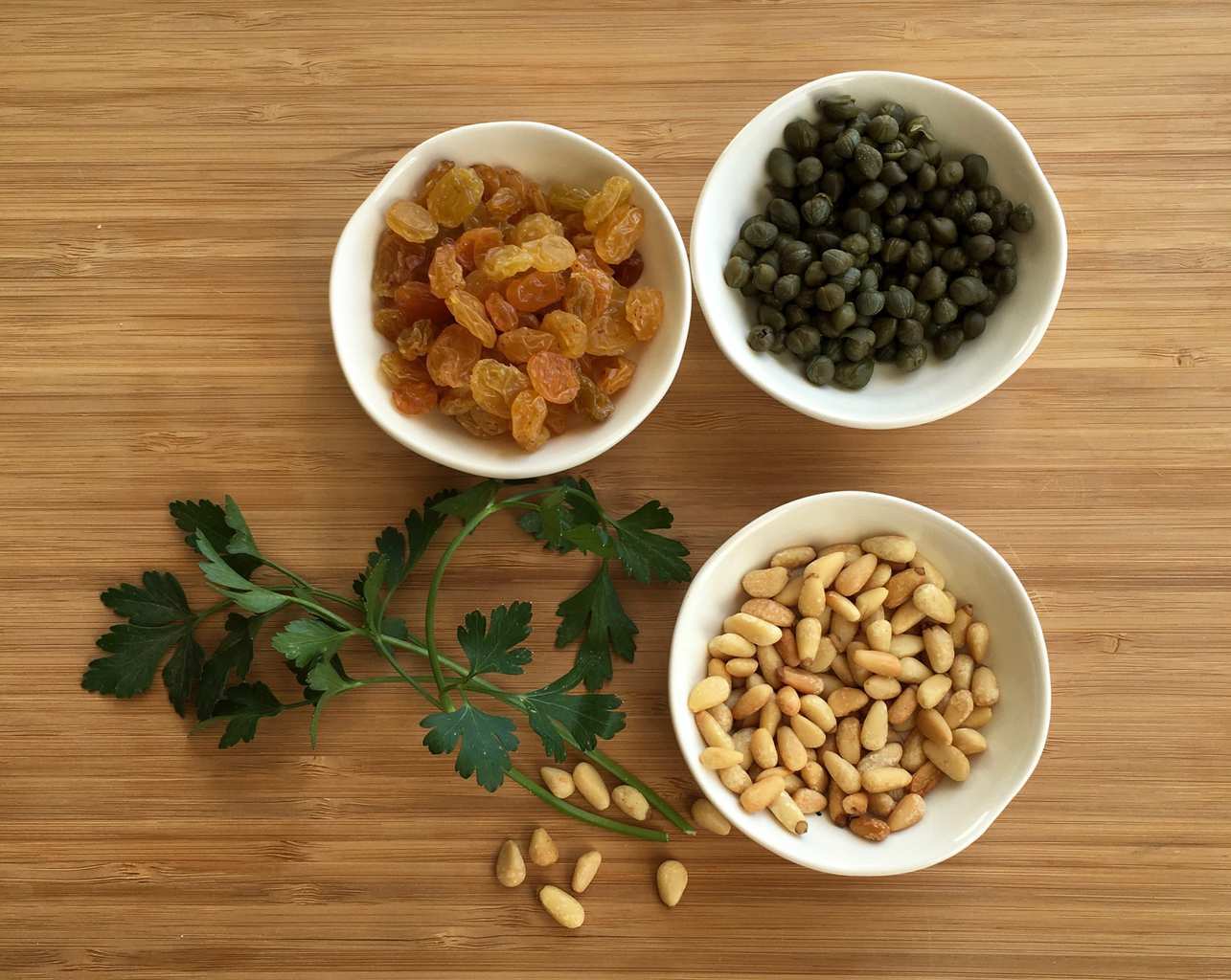 Overhead view of three small dishes: one of golden raisins, one of pine nuts, one of capers. 
