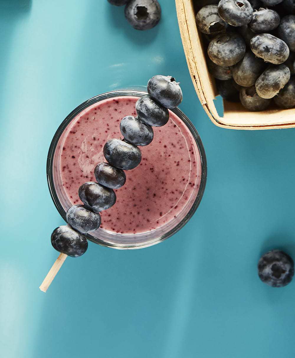 Blueberry Ginger Sizzle Smoothie with blueberry stir stick on turquoise background.