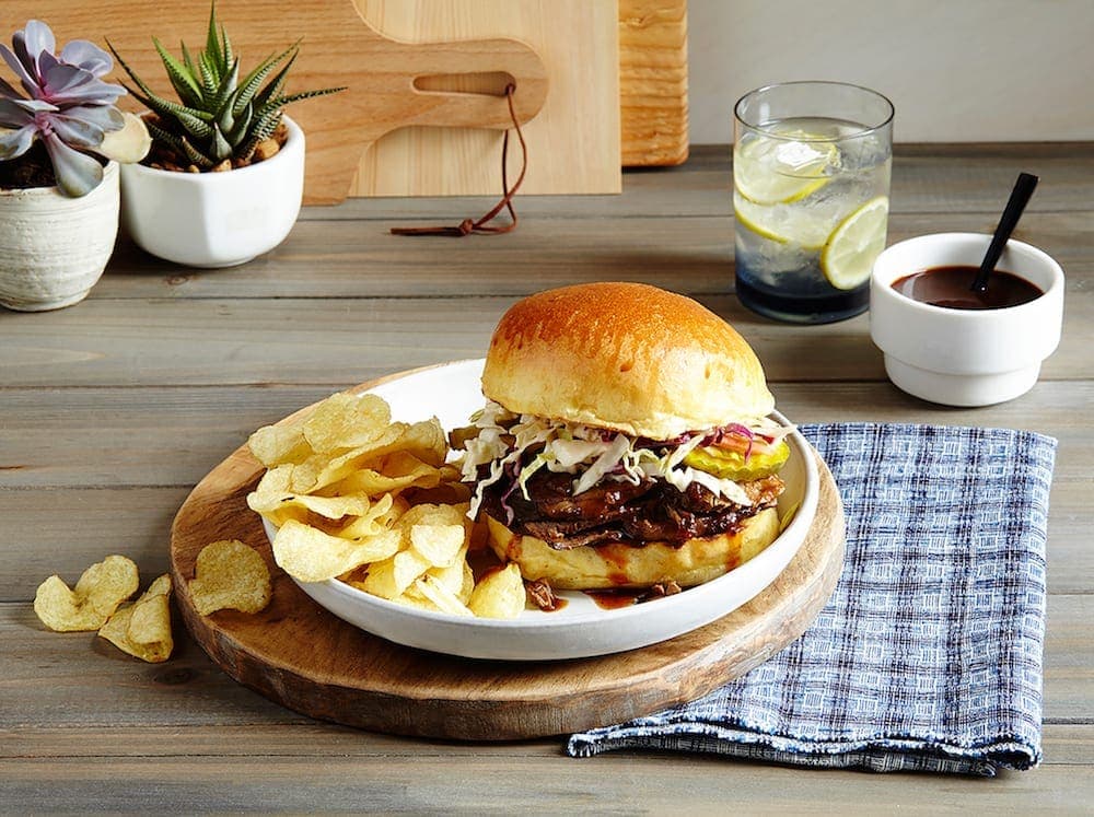 A Slow Cooker Brisket Sandwich on table, white plate and board, blue napkin.