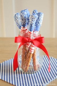 Red, white and blue dipped stick pretzels stand in a jar by an American Flag for easy 4th of July food shared by Tara Teaspoon.