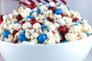 Caramel coated popcorn is mixed in with M&Ms, marshmallows ad sprinkles in a bowl with an American flag make great 4th of July food shared by Tara Teaspoon.