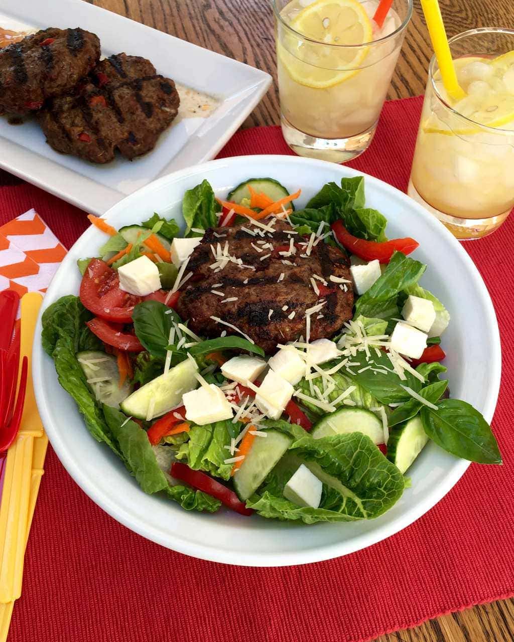 Grilled Italian bunless burgers in white bowl w red placemat and lemonade