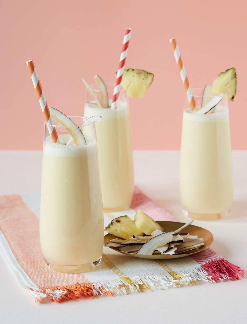 Three homemade pina coladas in tall glasses, garnished with straws, coconut strips, and pineapple chunks. 