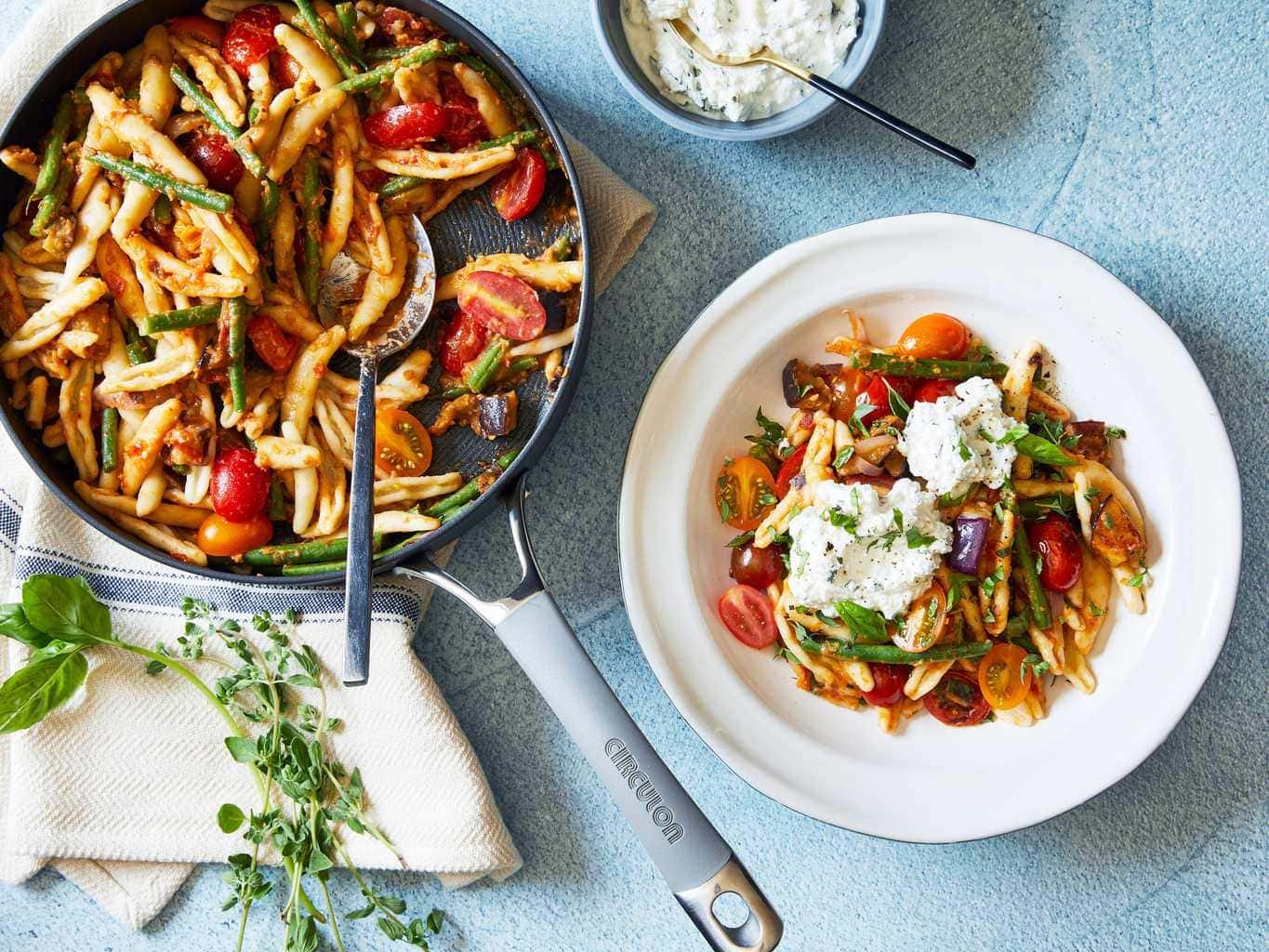 Italian tomato and eggplant skillet pasta in skillet and white bowl with ricotta on blue background