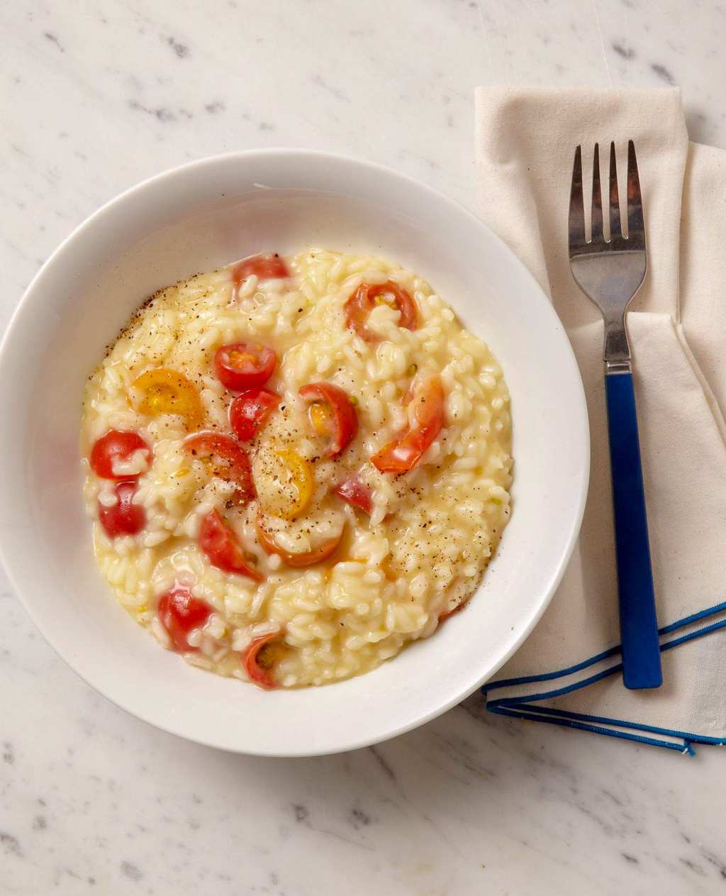 Creamy cherry tomato risotto with fresh tomatoes and cheese in white bowl w blue fork on marble surface
