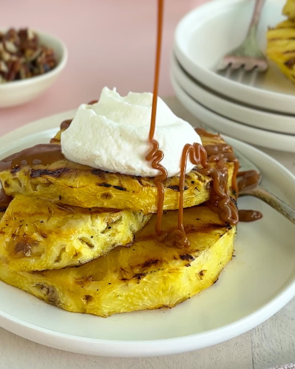 Grilled pineapple slices on a white plate topped with whipped cream and being drizzled with caramel.
