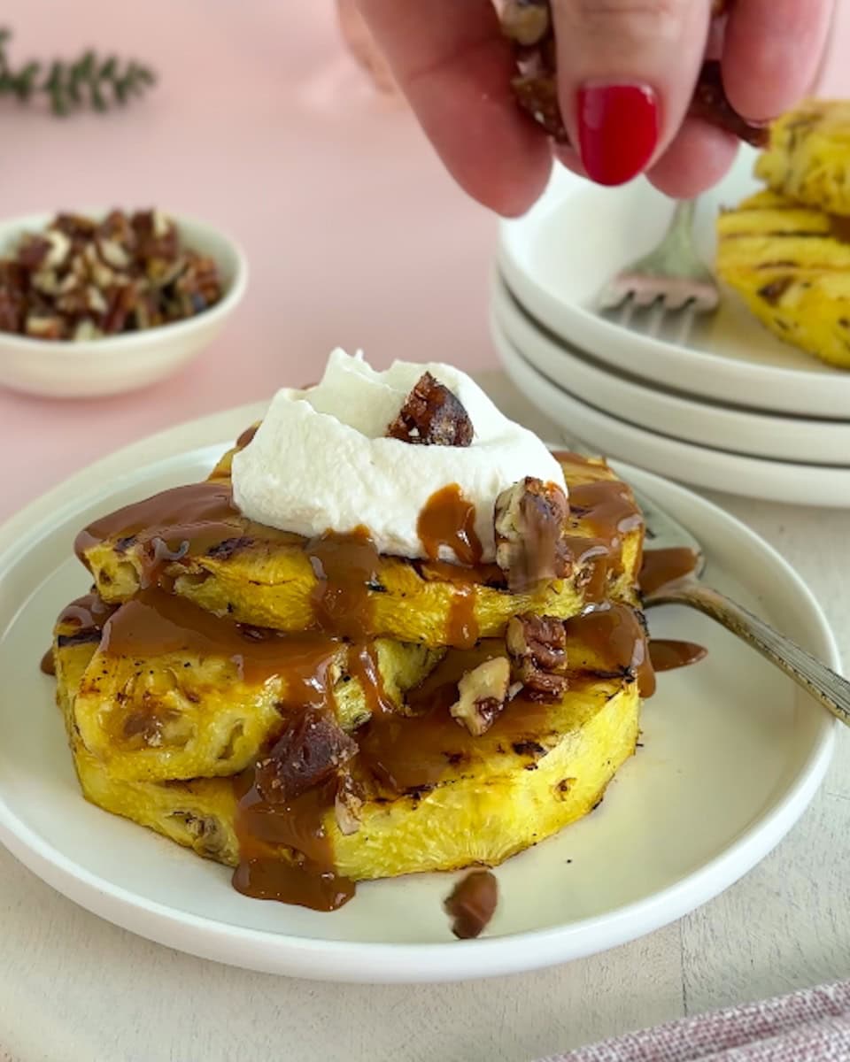 Grilled pineapple slices on a white plate topped with caramel, whipped cream, and pralines.