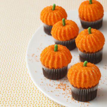 Homemade marshmallow pumpkins piped onto mini cupcakes for Halloween, with orange sugar sprinkles, on a serving platter.