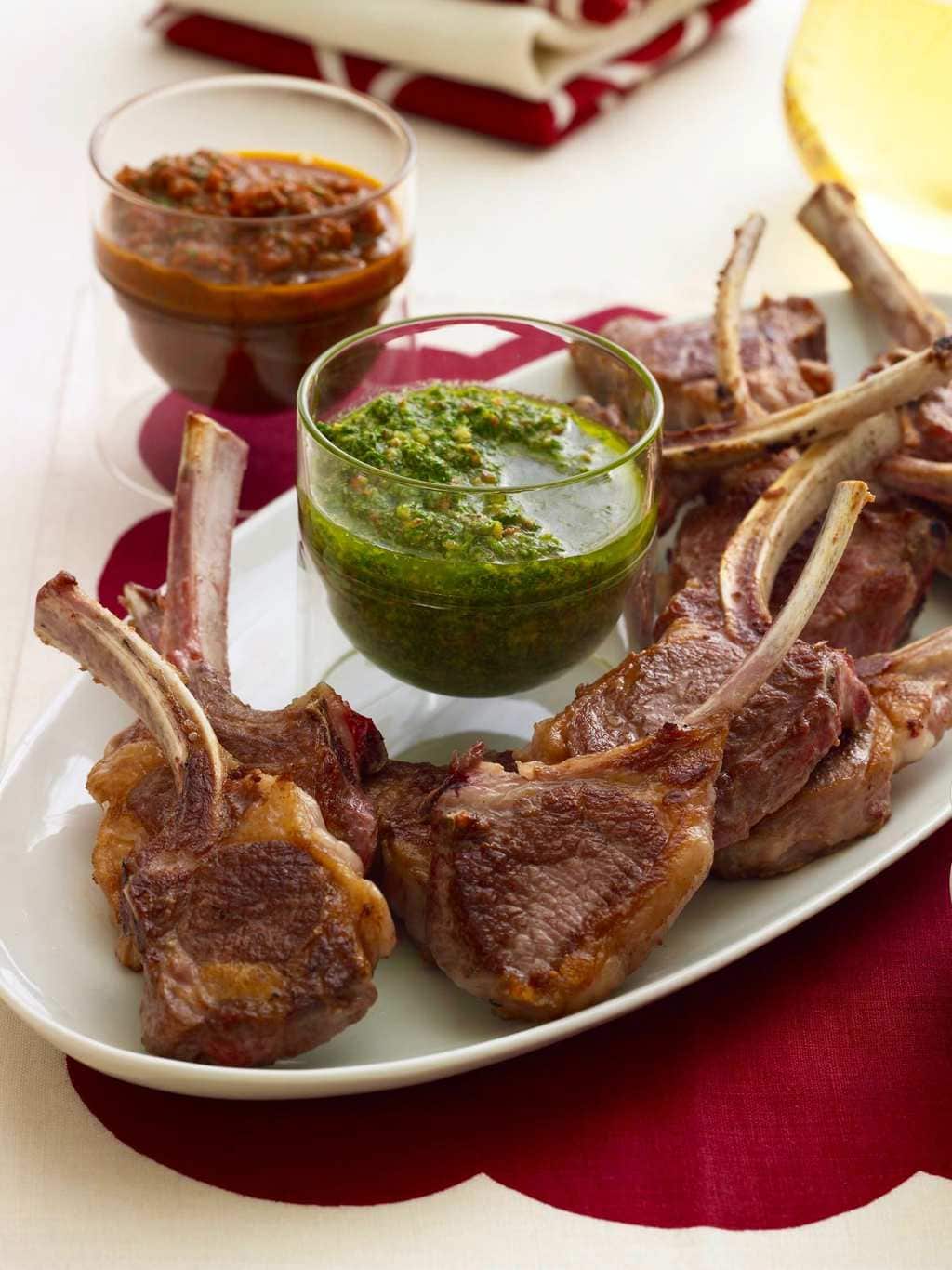 Baby lamb chops with red and green sauce on white oval platter, red linen