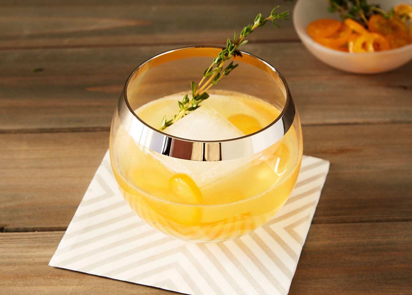 Kumquat Thyme Cocktail in gold-rimmed glass with thyme sprig on wood and striped napkin