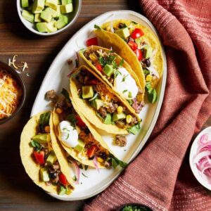 Chili Beef Tacos on white platter with avocado cubes and shredded cheese