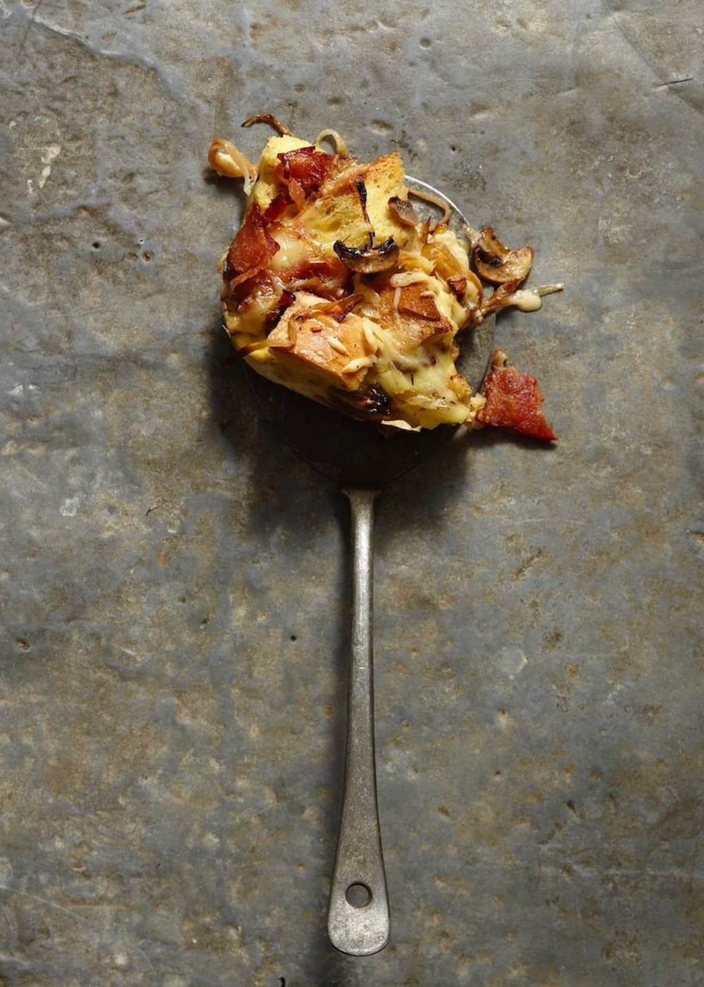 A serving of Onion Bacon mushroom strata on a metal serving spoon on stone background