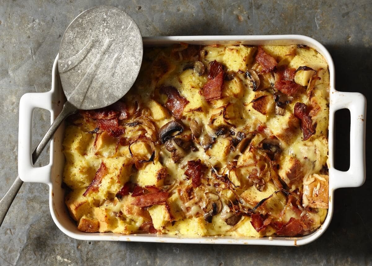 Baked Onion Bacon Mushroom Strata in white casserole dish with metal serving spoon