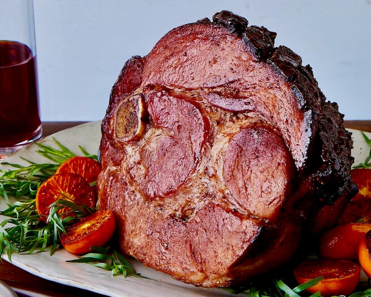 Roast ham with spiced honey glaze on white platter surrounded by fresh herbs and roasted tomatoes.