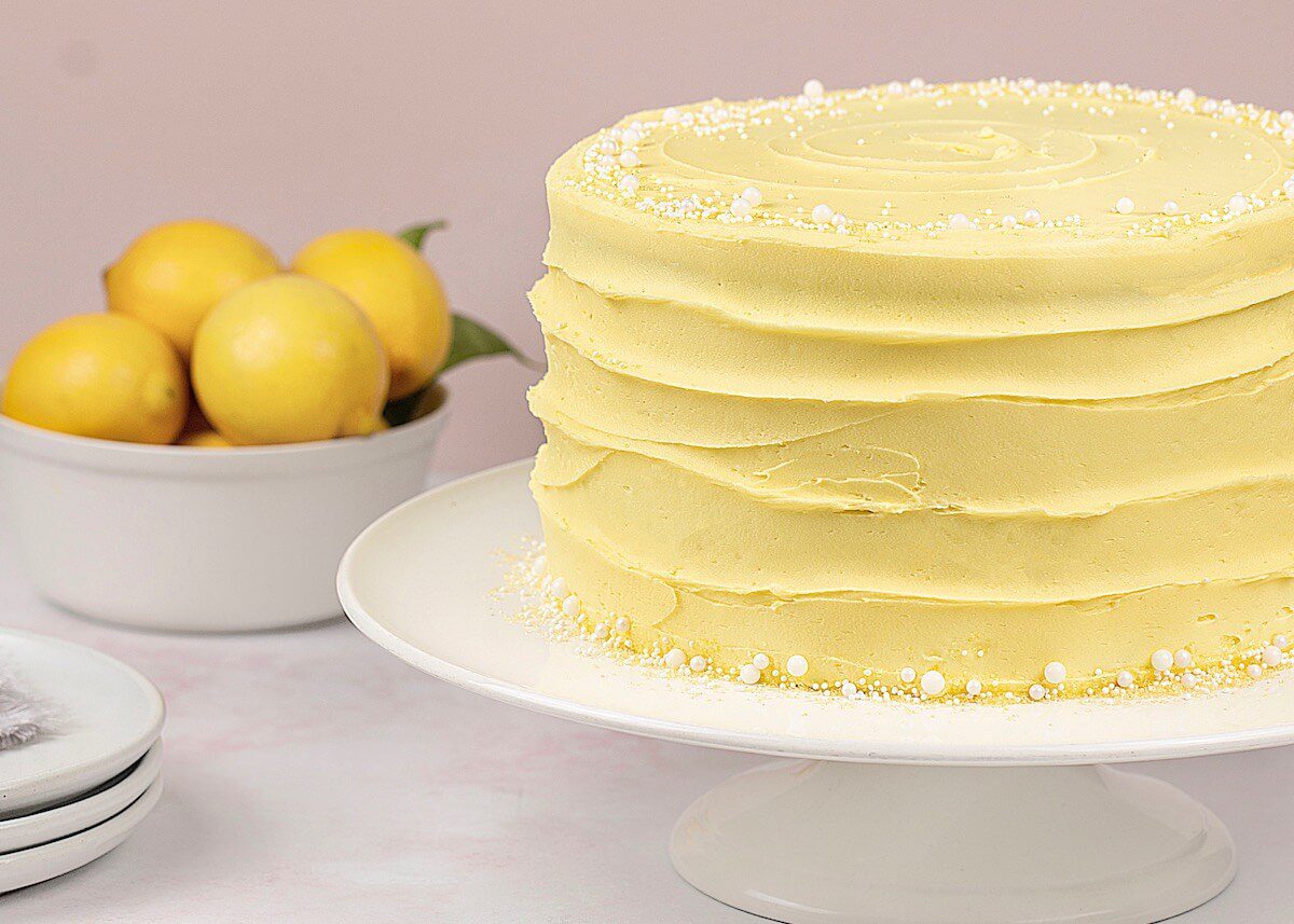 Lemon Cake on white cake stand with a bowl of fresh lemons in the background