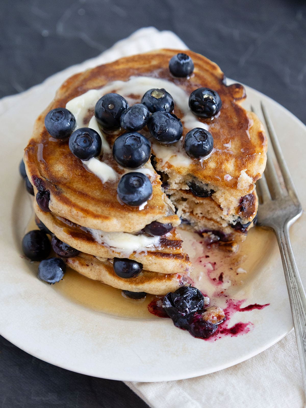 A stack of blueberry pancakes topped with butter, syrup and fresh berries with a large bite removed. 