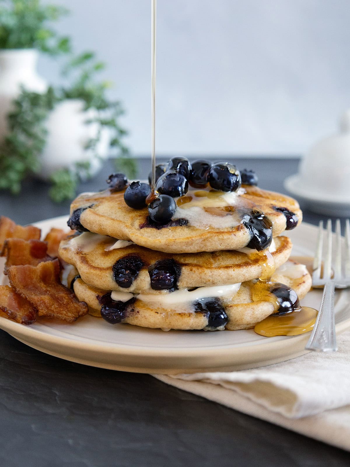 A stack of blueberry pancakes topped with butter and fresh berries being drizzled with syrup.