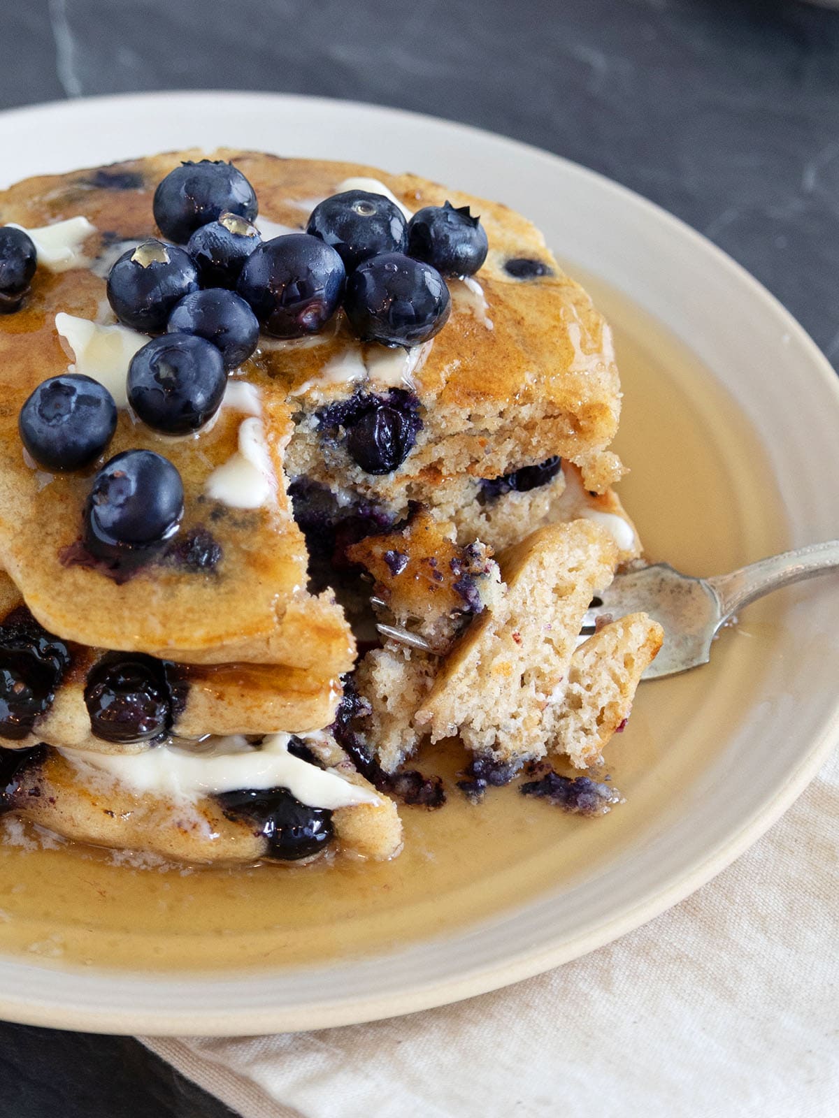 A stack of blueberry pancakes topped with butter, syrup and fresh berries with a large bite on a fork.