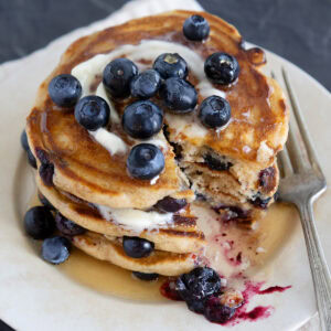 A stack of blueberry pancakes topped with butter, syrup and fresh berries with a large bite removed.