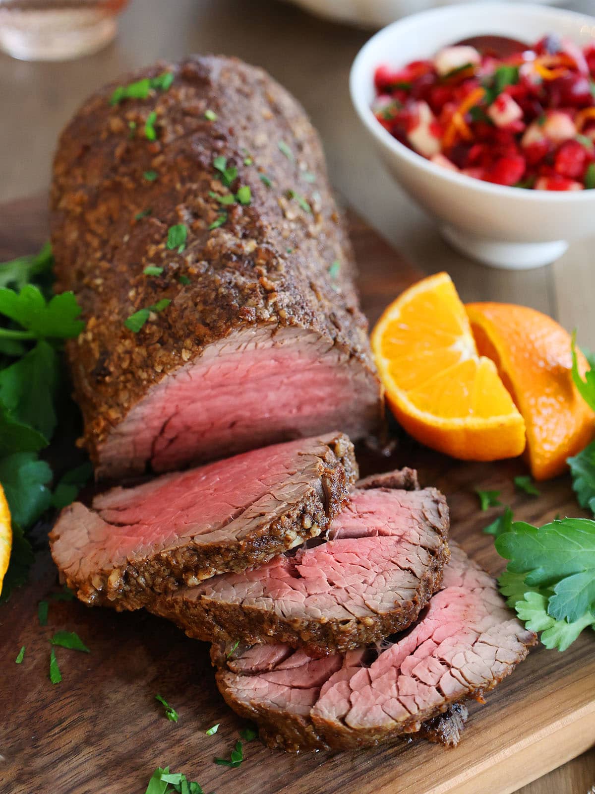 Close up of a beef tenderloin roast on a cutting board next to oranges and a bowl of cranberry relish. 