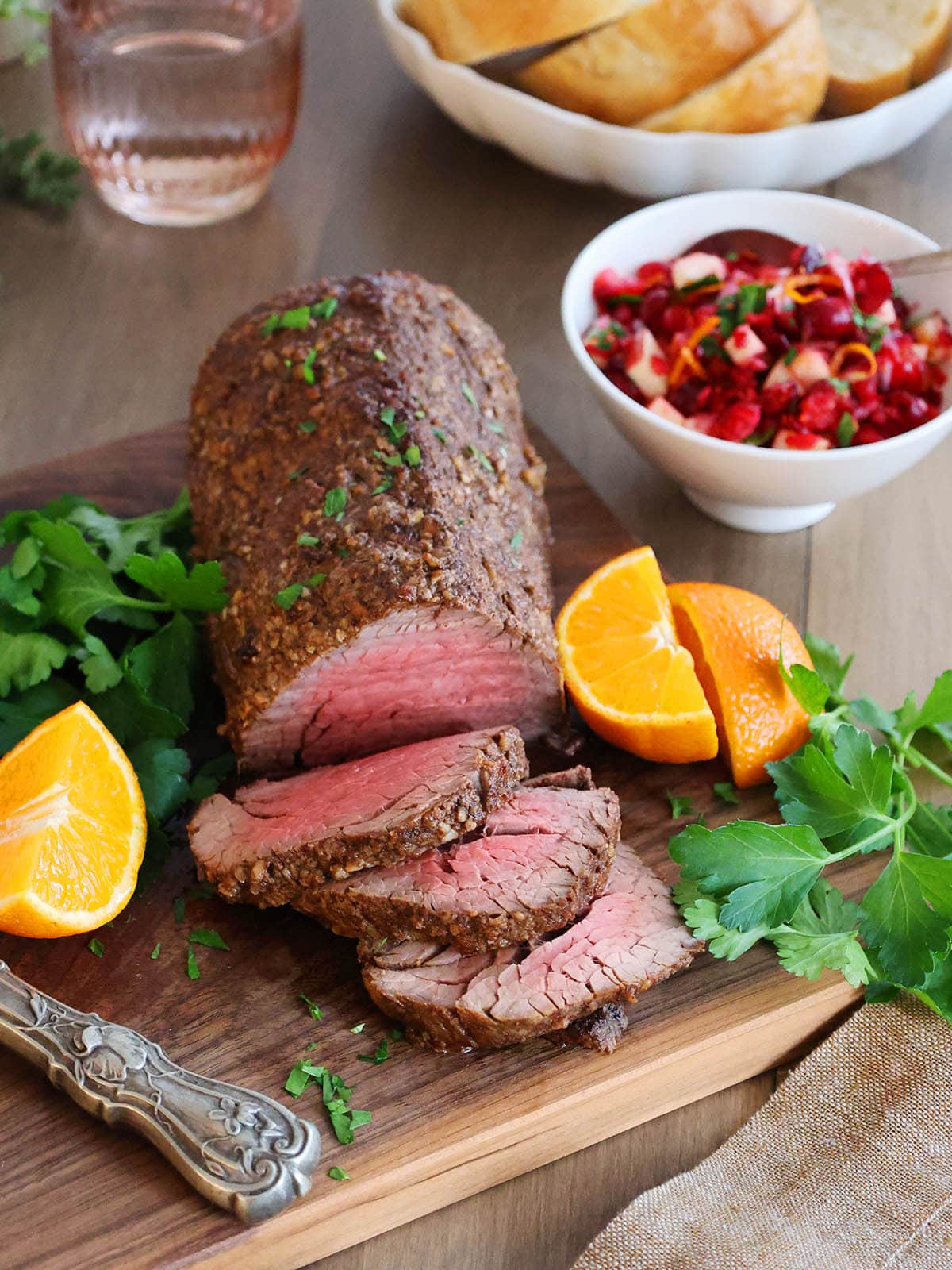 A beef tenderloin roast on a cutting board next to oranges, parsley and a bowl of cranberry relish. 