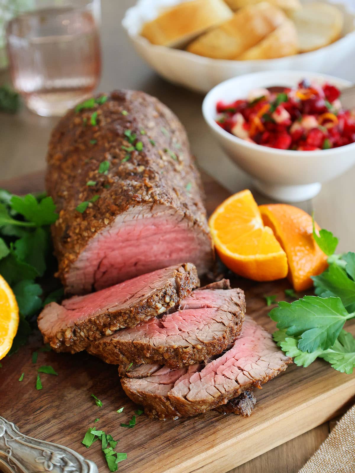 A beef tenderloin roast with a few slices removed on a cutting board next to oranges and a bowl of cranberry relish. 