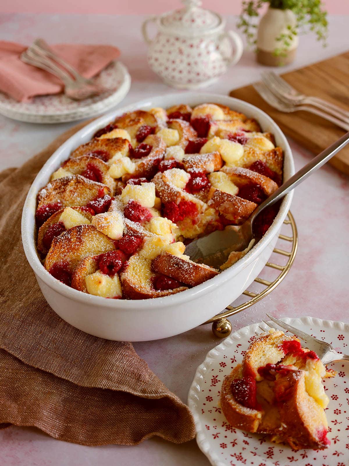 Raspberry french toast casserole in a white baking dish with a serving removed on a small white plate.