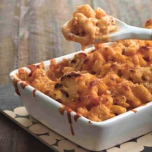A spoon holding up a serving of cauliflower mac and cheese over the baking dish.