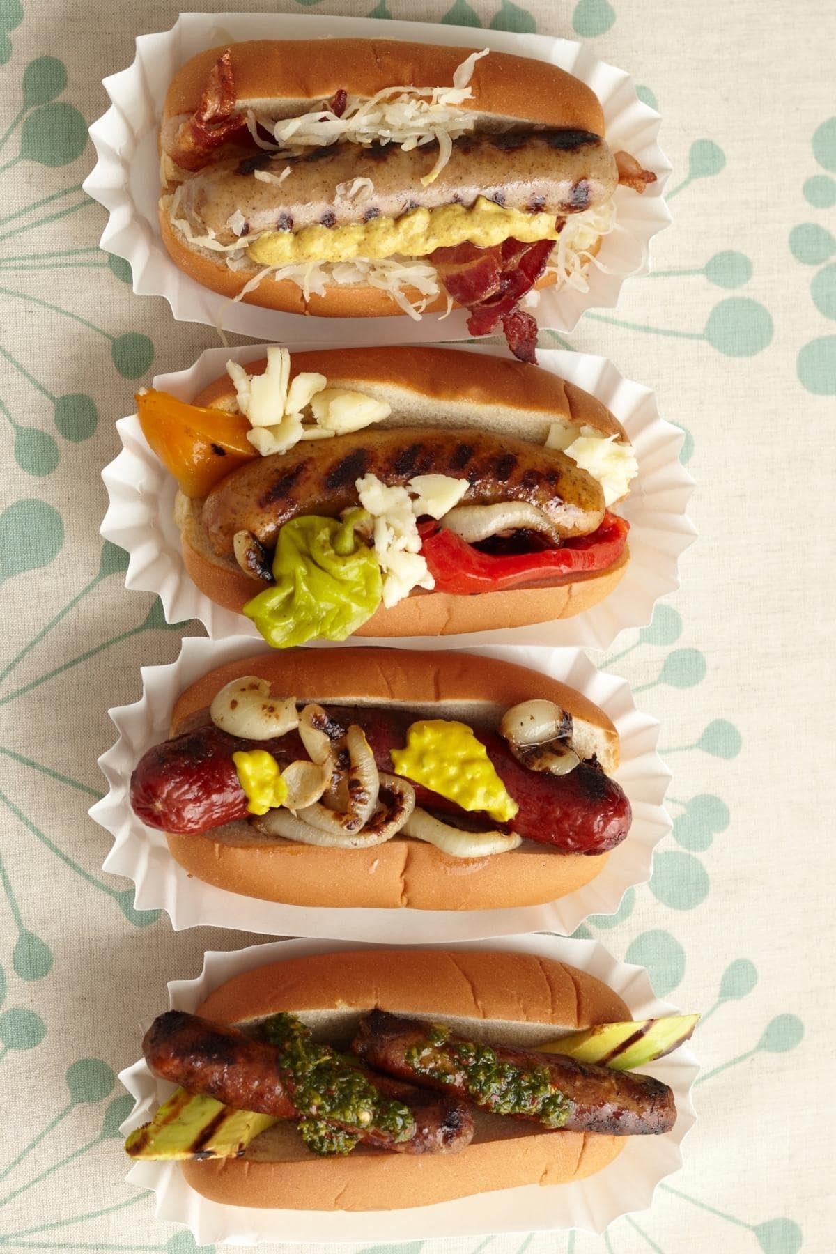 Four gourmet hot dogs in a row on a table cloth.
