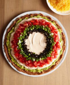 Adding the center cheese layer to a beautiful 7 layer dip on a round white platter.