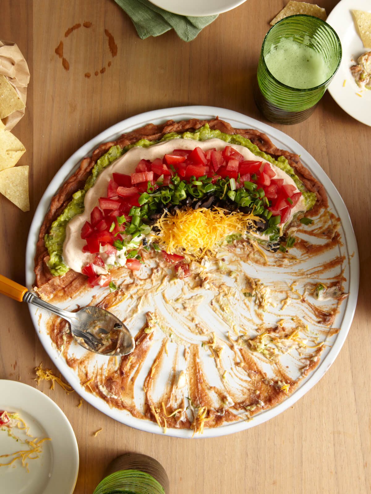 7 layer dip half eaten on a white round platter on wooden table party scene.