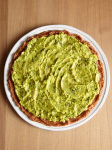 Guacamole spread over bean dip on a white platter making 7-layer dip.