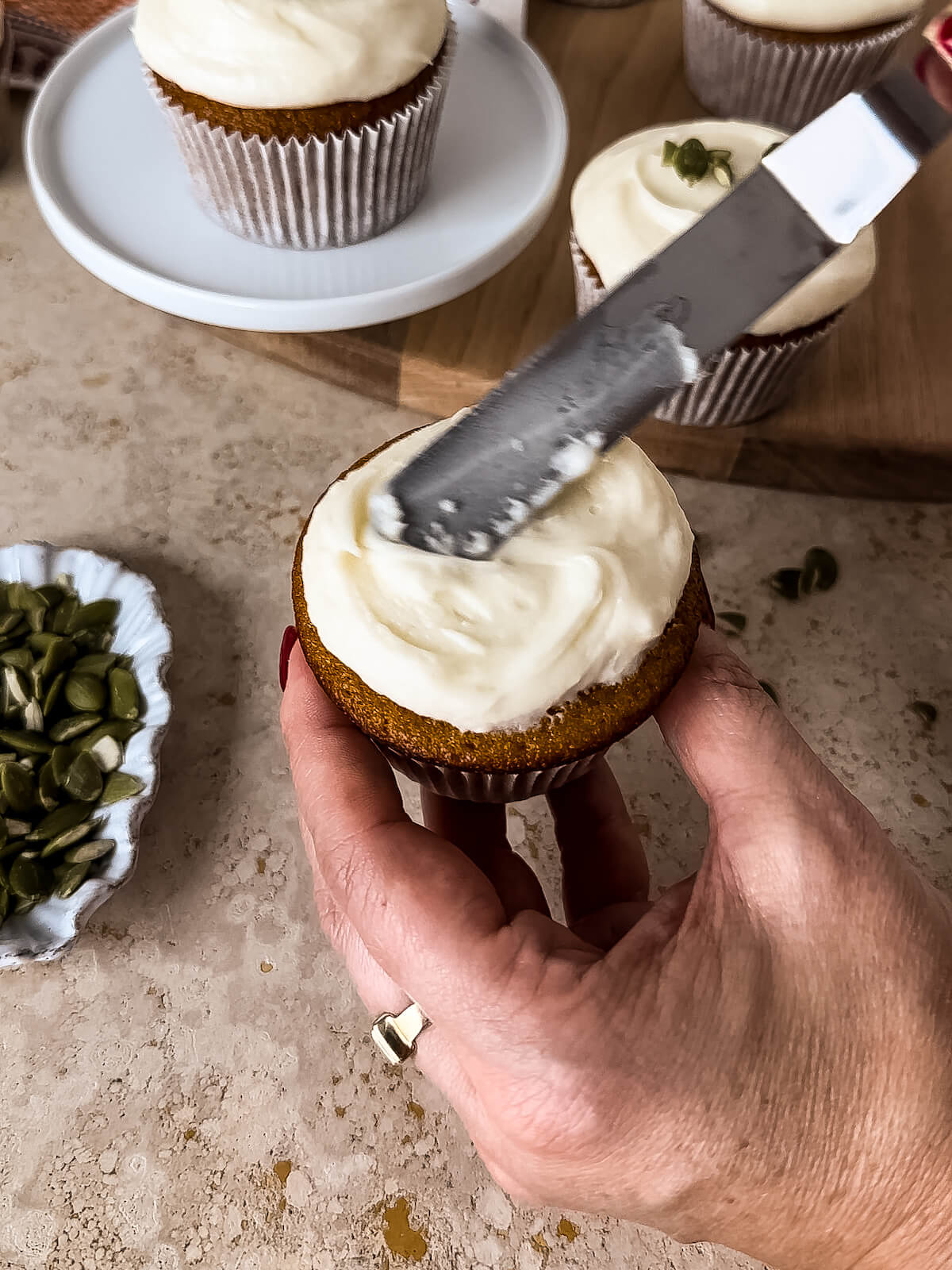 Hand holding pumpkin cupcake, frosting it with cream cheese frosting using an offset spatula.
