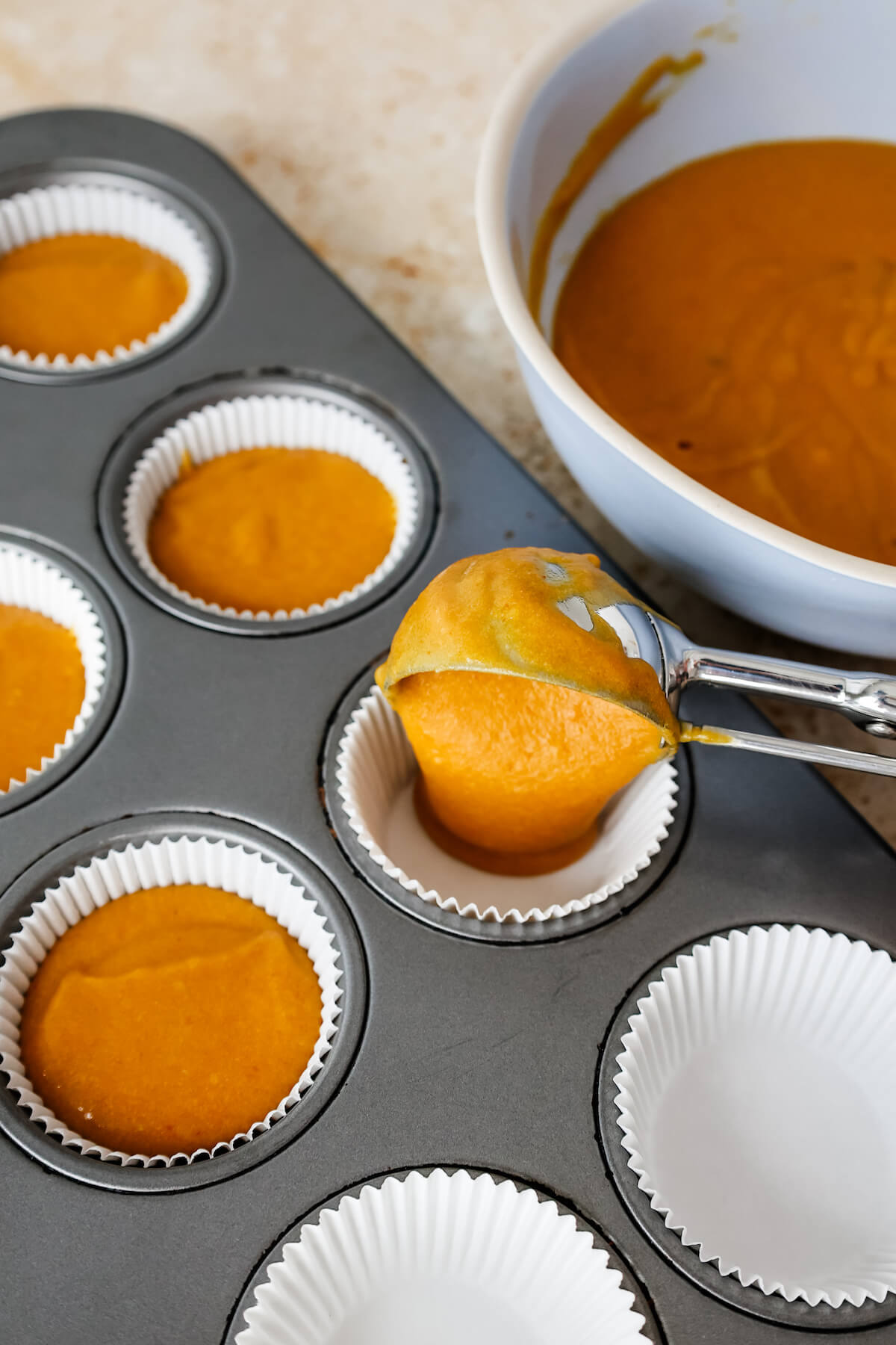 Cookie scoop portioning orange pumpkin cupcake batter into white muffin cups in a muffin tin.