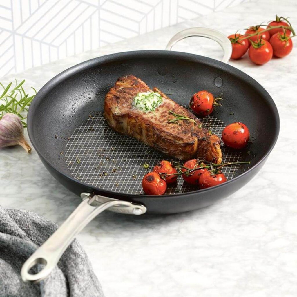 Photo of the 12-in Analon Hybrid Nonstick Frying Pan as it cooks a well-seared stake and tomatoes on a marble counter top.