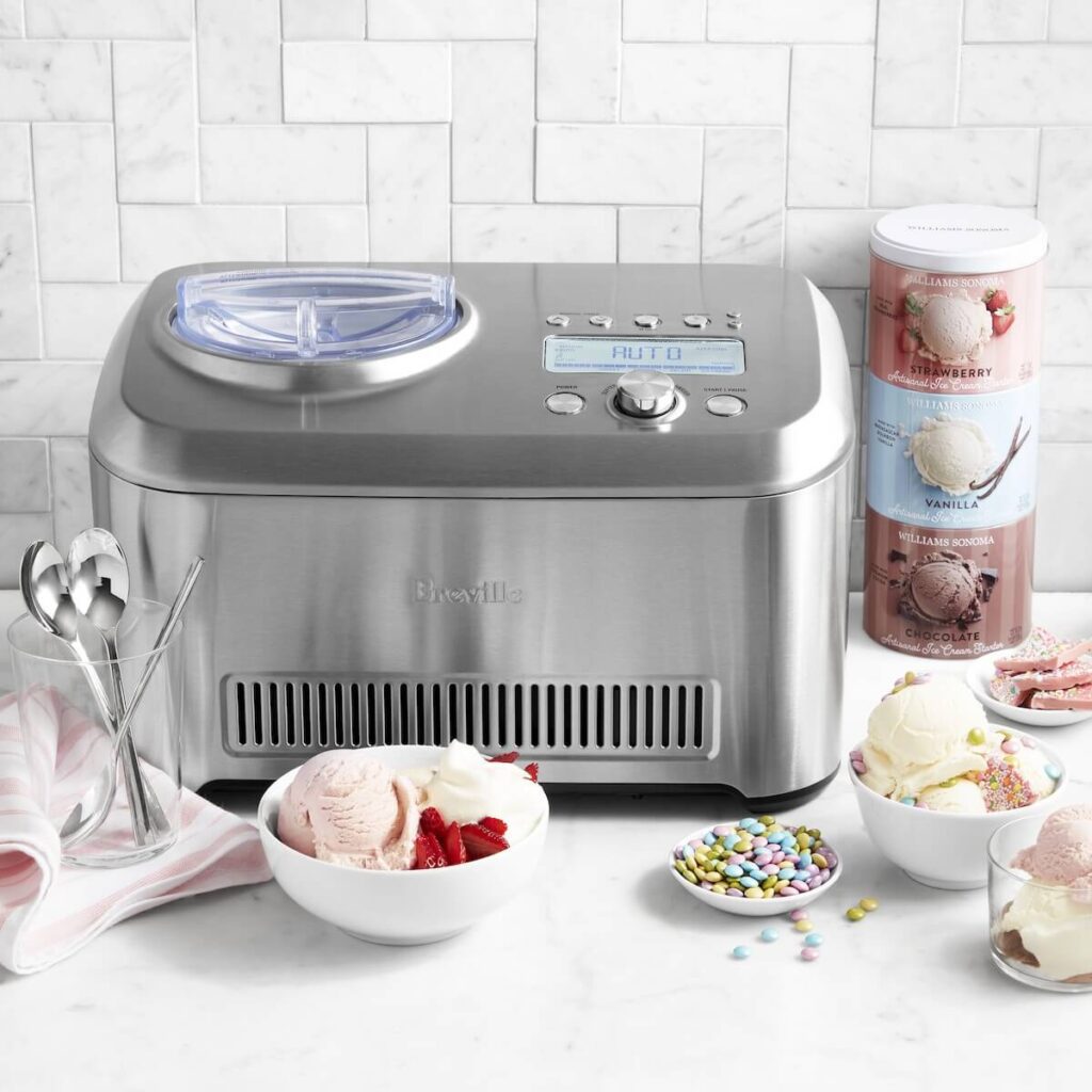The Breville smart scoop ice cream maker in brushed stainless steel set in a white kitchen with ice cream mixes behind it and already made vanilla and straberry ice cream in front, scooped into white bowls and topped with mini m&ms.
