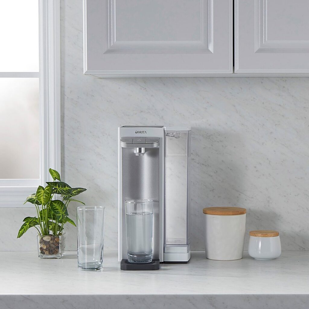 12 cup Brita Hub Countertop Water Filter System in white place on a white kitchen counter with a cup under its spout filled with clean water.