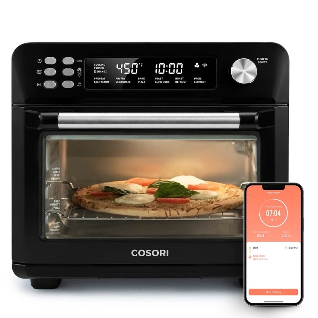 A black Cosori Smart Air Fryer Toaster baking a pizza with tomatoes and mozzarella, with a phone connected to it displaying its wireless control functionality.