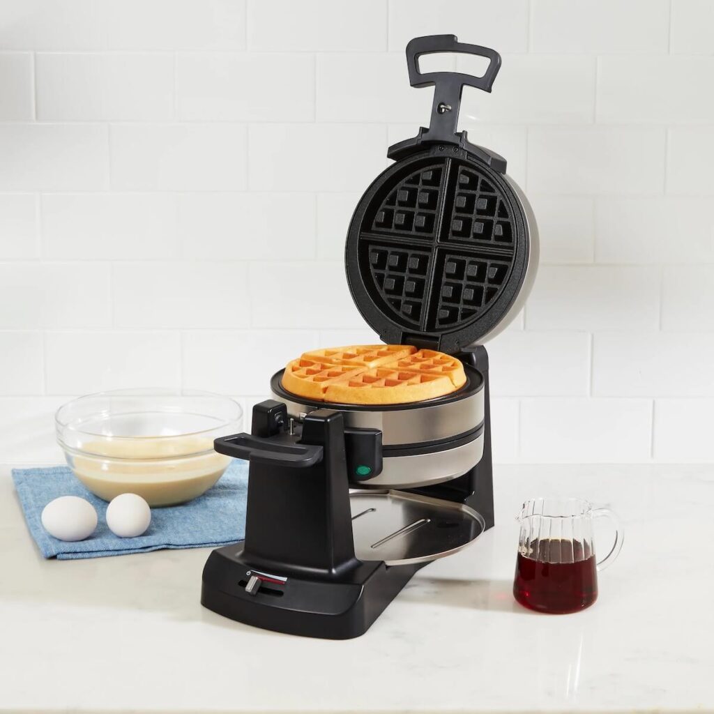 A double flip Cuisinart waffle maker cooking a waffle, set in a white kitchen with waffle batter and maple syrup close by.