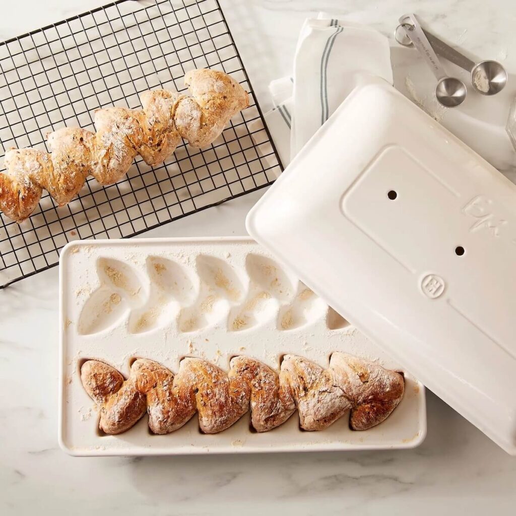 Emile Henry's Epi Bread baking pan in color linen, placed in a white kitchen with two loaves of bread freshly baked, on still placed in the pan and one cooling on a wire rack.