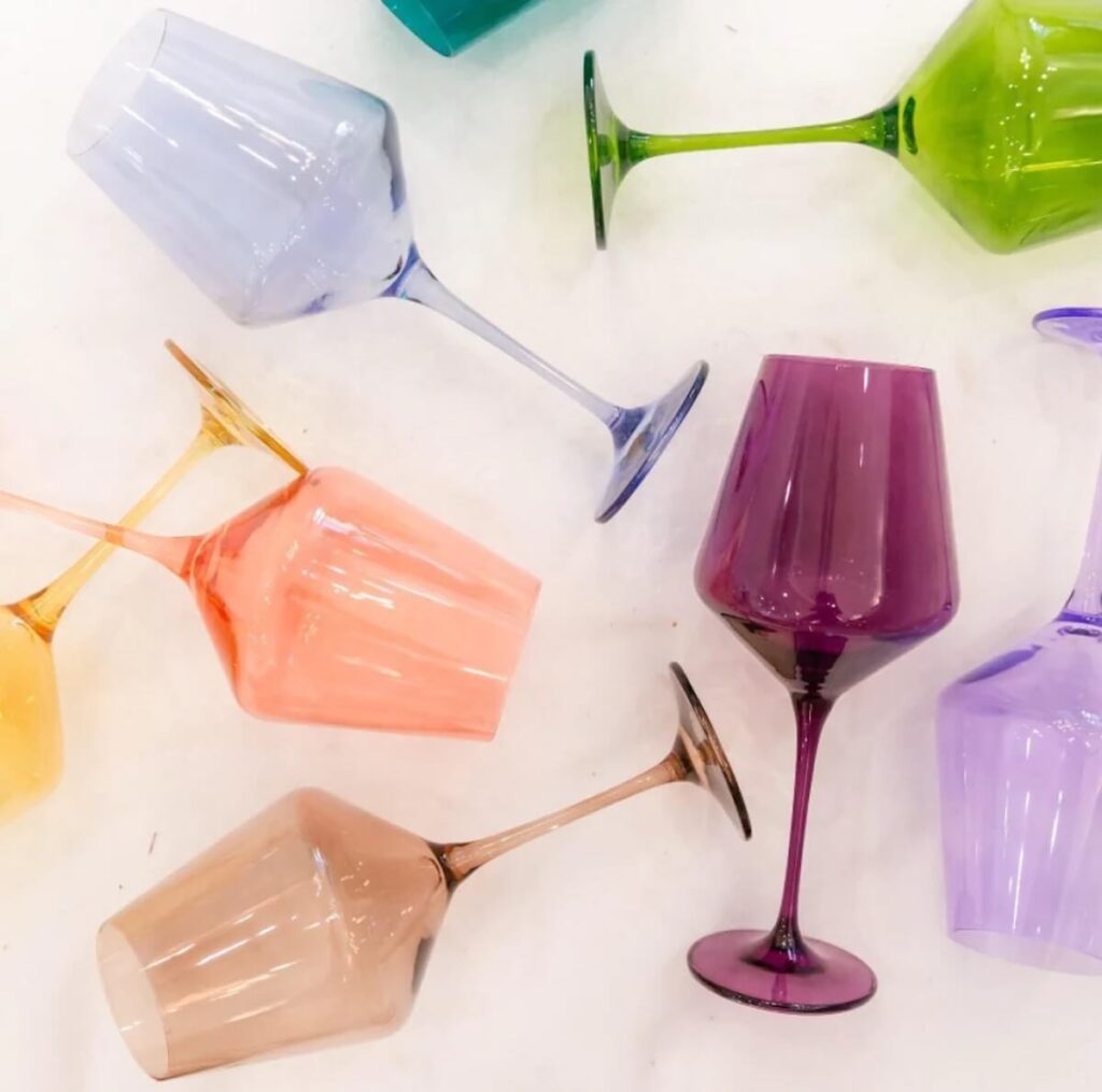 Multiple colors of estelle's colored wine glass collection laid out over a white surface, including dark purple, bright gree, light purple, peach, yellow. brown, and more.