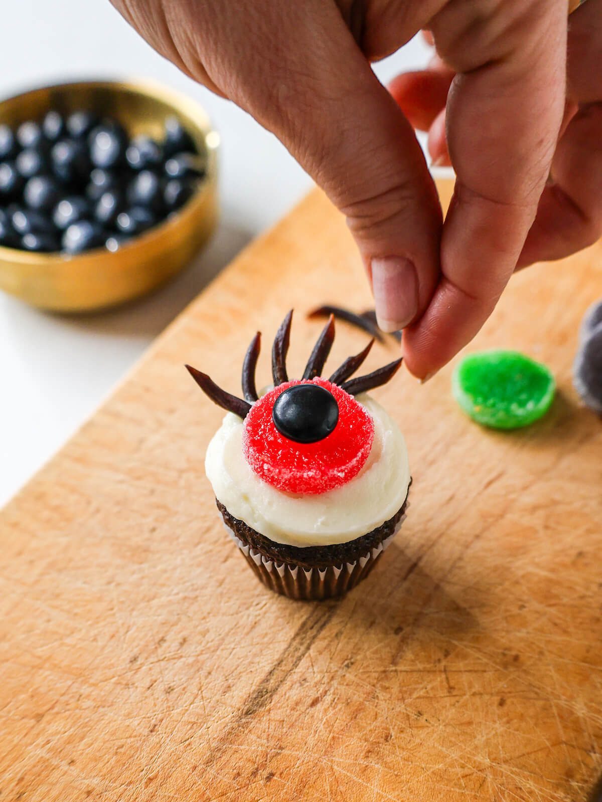 Finishing adding cut licorice laces on a white frosted cupcake with a candy eyeball to create eyelashes.
