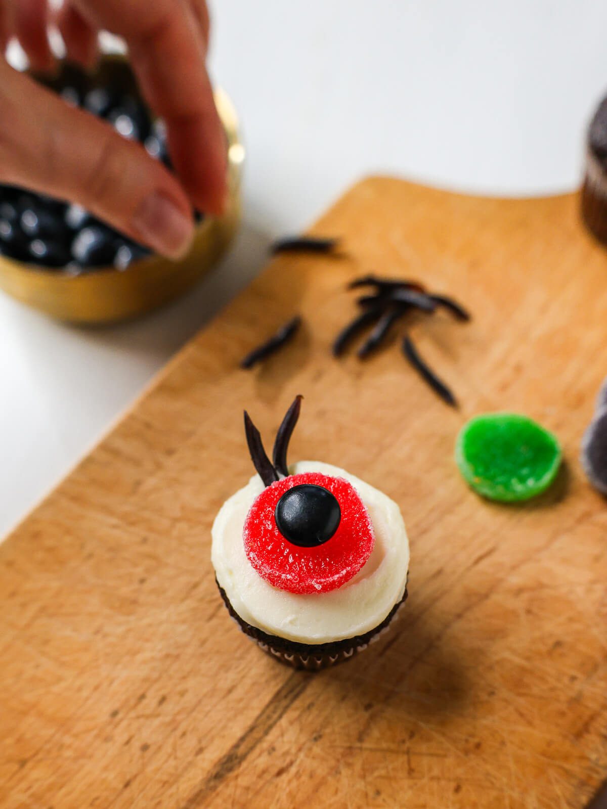 Placing cut licorice laces on a white frosted cupcake with a candy eyeball to create eyelashes.