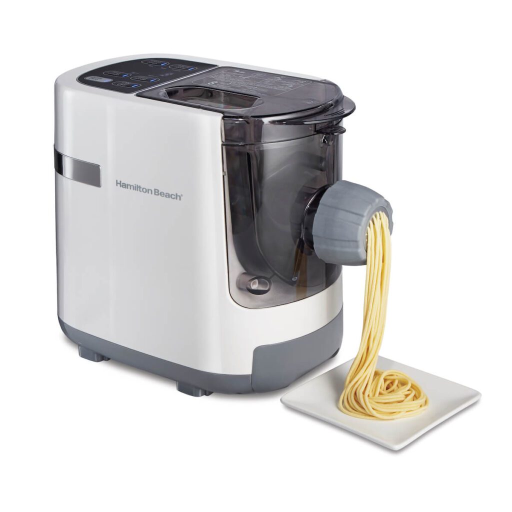 A White hamilton beach pasta maker over a while background, dispensing freshly made spaghetti noodles from its spout.